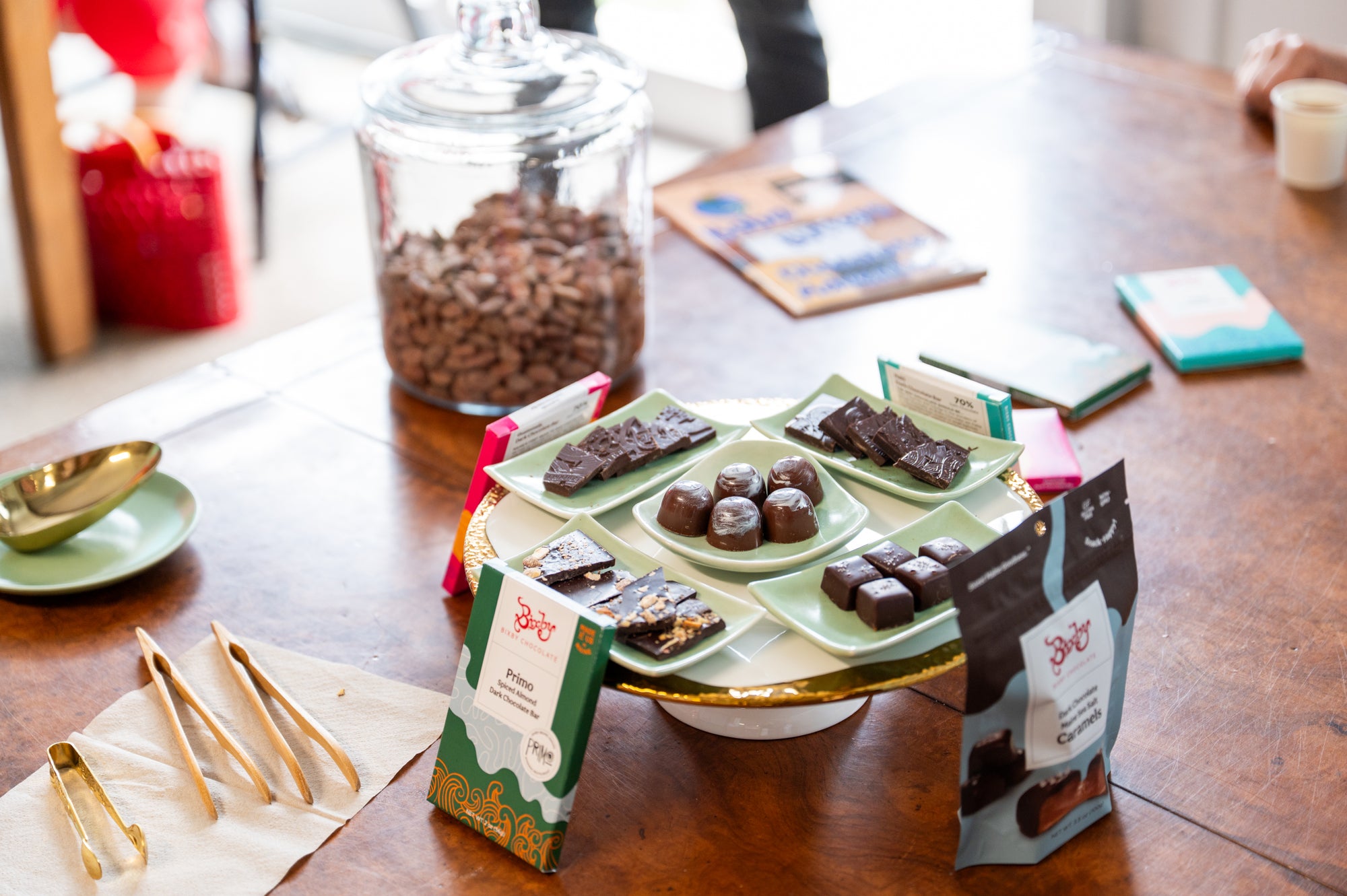 A display of assorted chocolates on green plates sits on a wooden table with packaged chocolate bars, bags, gold tongs, a glass jar of cocoa beans, and books in the background.