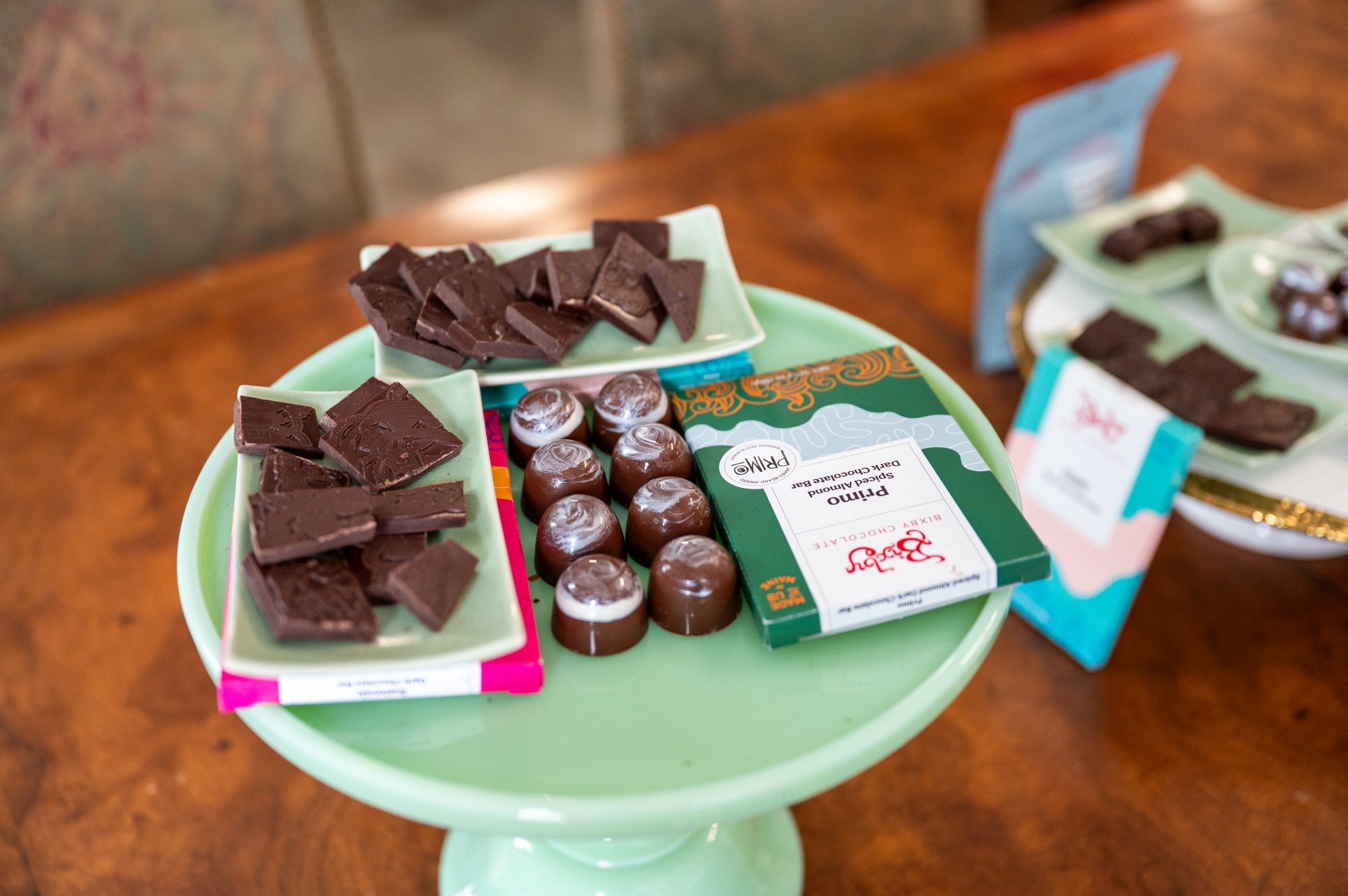 A green cake stand displays assorted dark chocolate squares, wrapped chocolate bars, and round chocolate truffles, with another tray of chocolates blurred in the background on a wooden table.