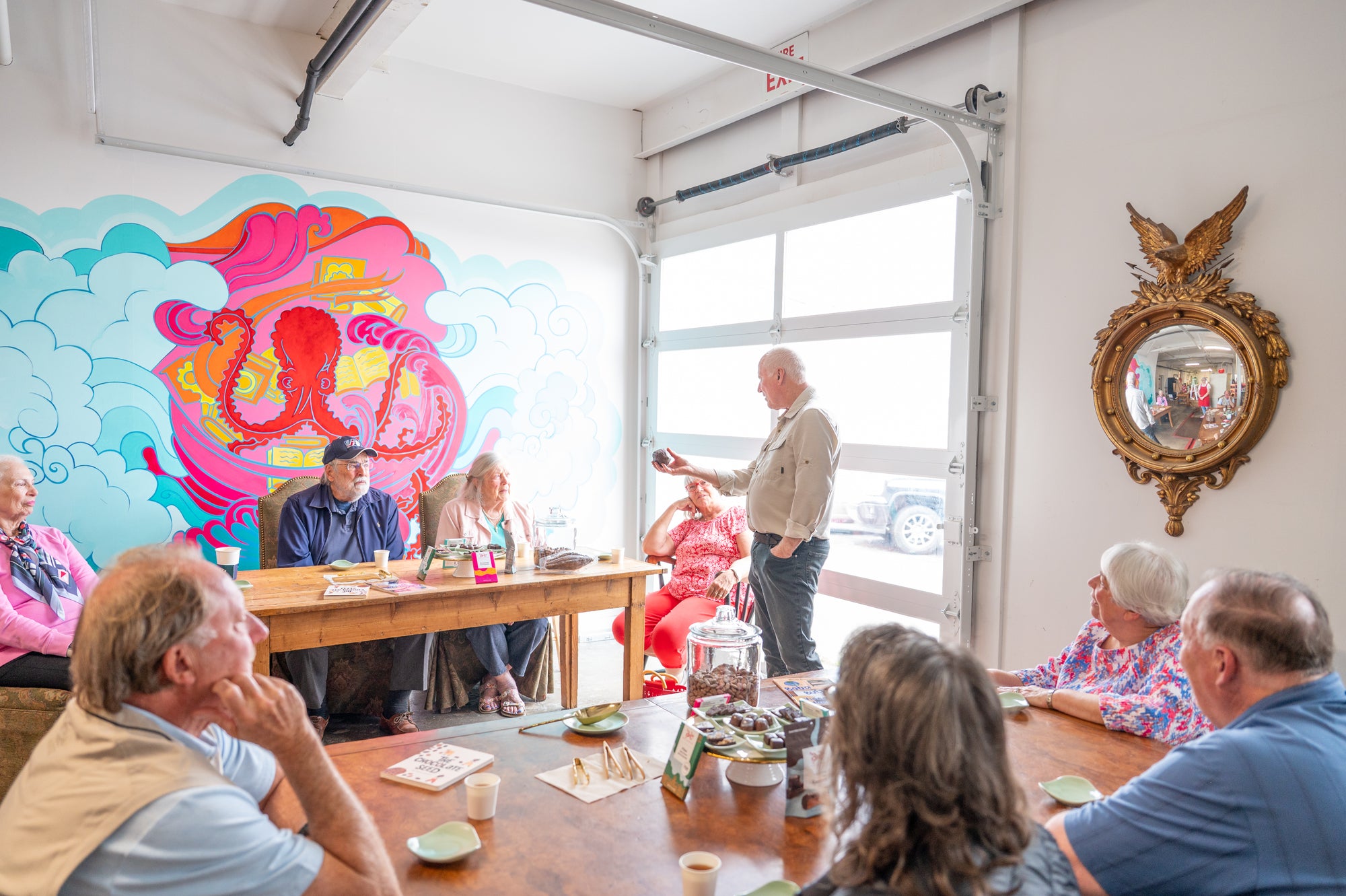 A group of older adults sit around tables in a bright room, listening to a man standing and speaking. Colorful wall art and a decorative mirror are visible in the background. Snacks and drinks are on the tables.