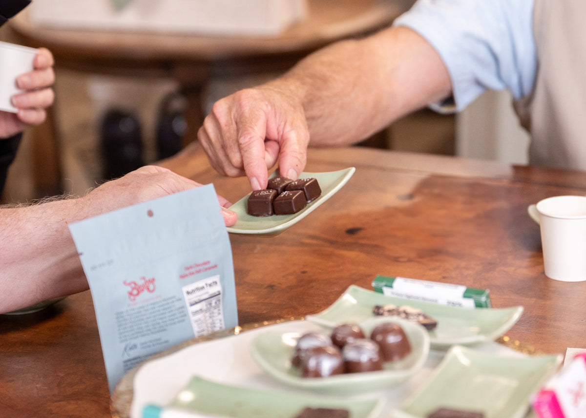 Two people reach for dark chocolates from a plate, savoring the Bixby Chocolate Tasting Experience featuring assorted treats and bean-to-bar chocolate inspired by Bixby Chocolate.