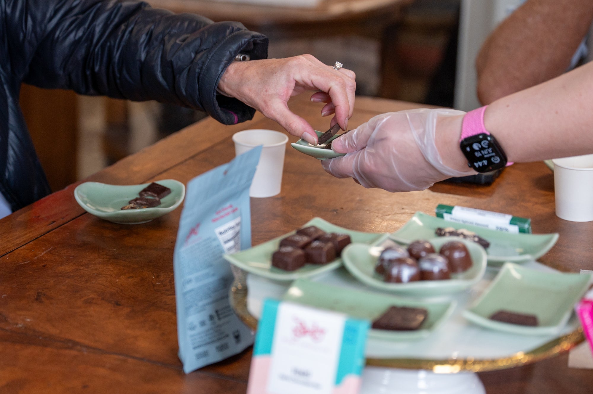 A person wearing a pink smartwatch and gloves hands chocolates on a green plate to another person across a wooden table with assorted chocolates and snacks.