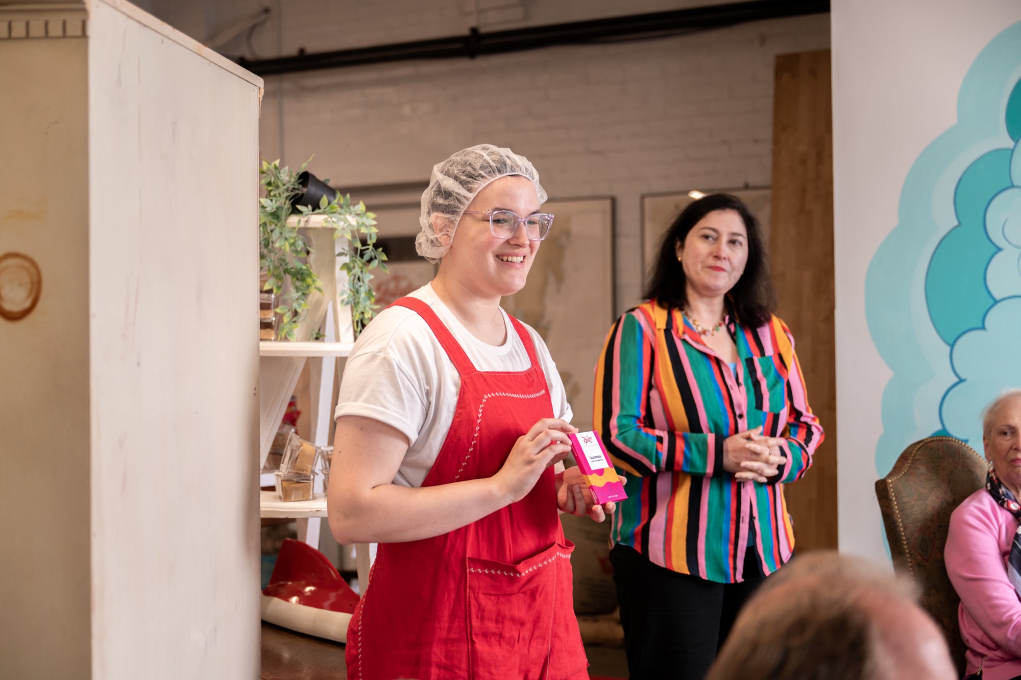 A person in red overalls and a hairnet holds a pink item, smiling, while another person in a colorful striped shirt stands nearby. They are indoors, with other people and decorations visible in the background.