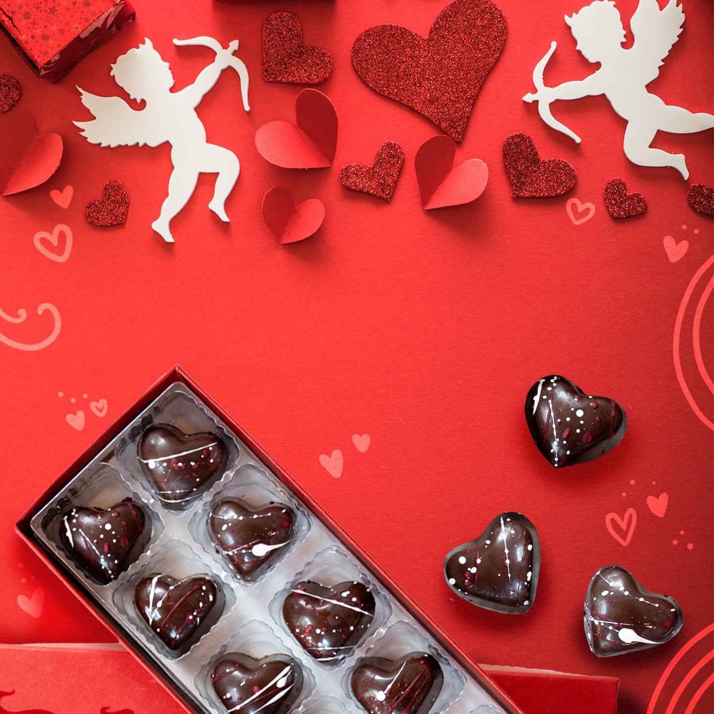 A box of heart-shaped chocolates sits on a red background decorated with paper hearts and cupids, creating a festive and romantic Valentines Day theme.