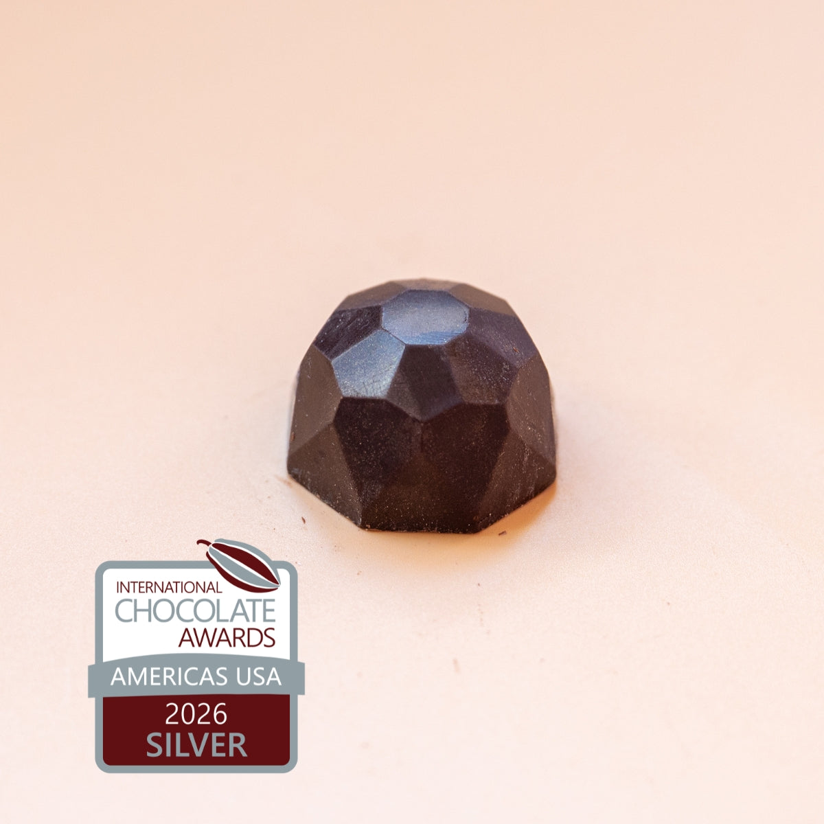 A single faceted dark chocolate truffle on a warm beige background, with an International Chocolate Awards 2026 Silver badge in the lower-left corner.