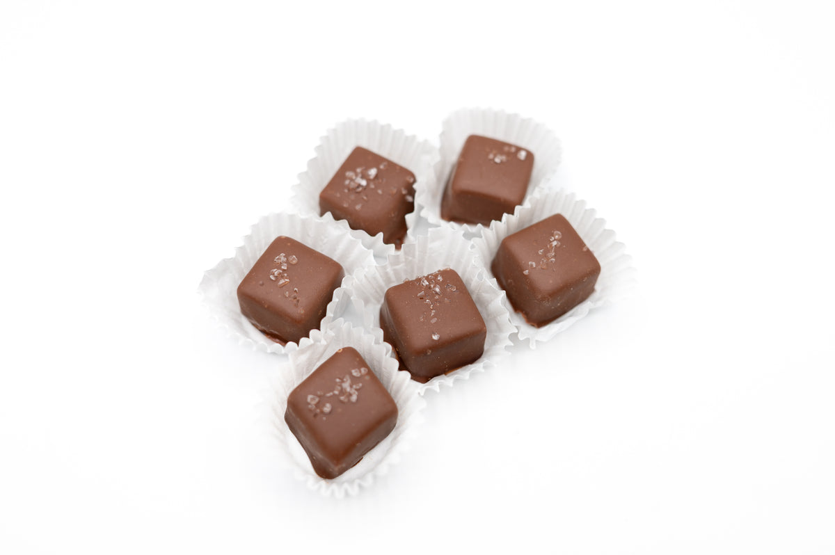 Six artisanal Chocolate Sea Salted Caramels from Bixby Chocolates are arranged in white paper cups, each topped with sea salt—making a perfect gift box for any occasion.