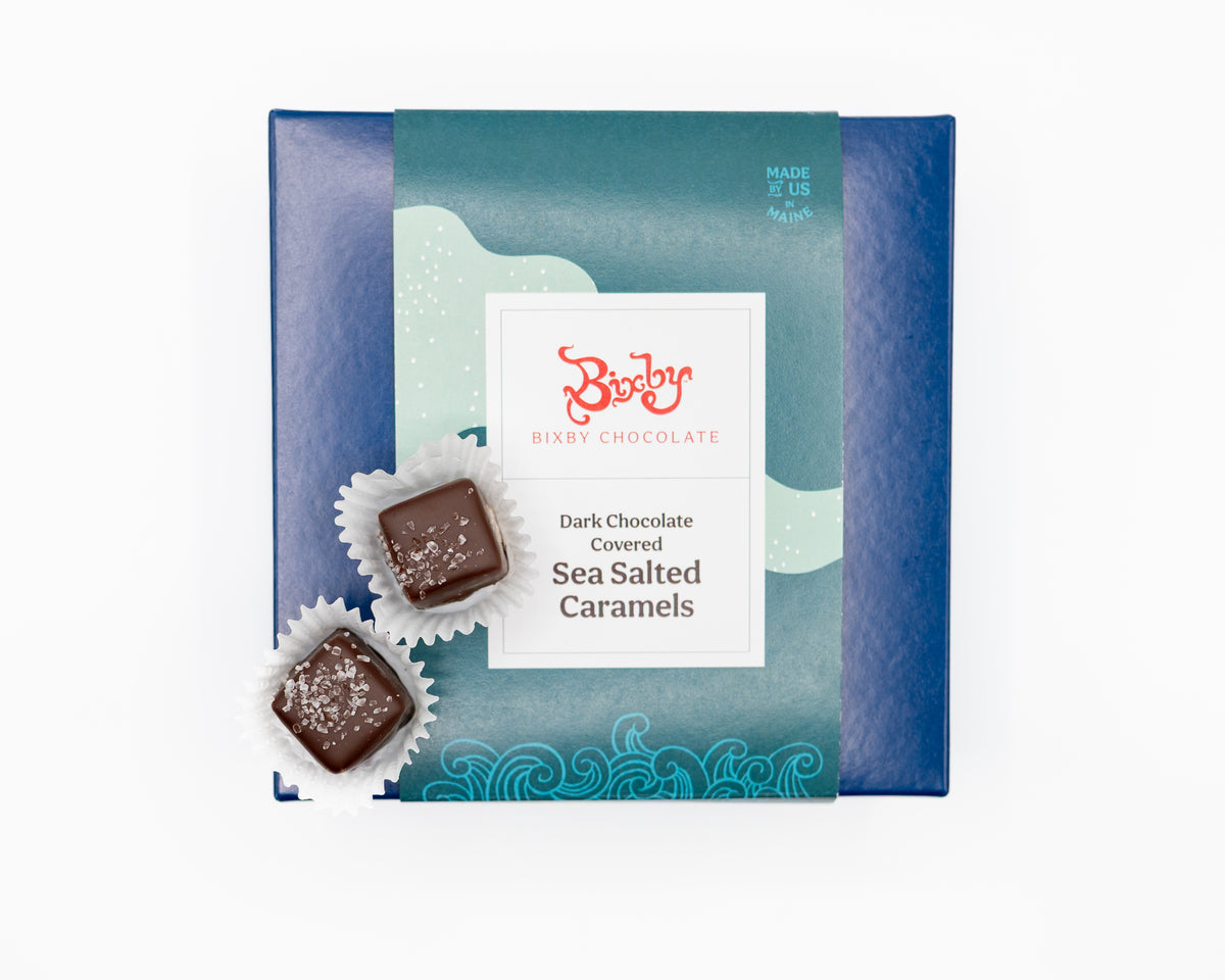 A blue box of Bixby Chocolates Chocolate Sea Salted Caramels Gift Box sits on a white background, with two unwrapped sea salt-topped dark chocolate caramels in front.