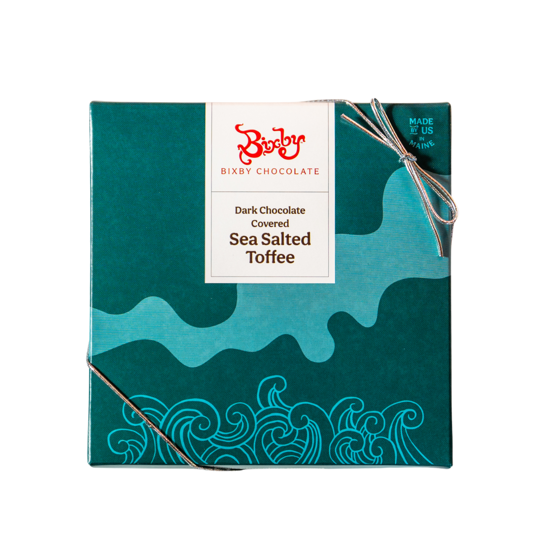 A teal box of Bixby Chocolates Dark Chocolate Toffee, featuring ocean wave designs, a white label, and the red Bixby logo, is tied with a silver string.