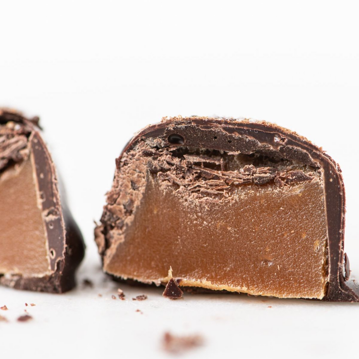 A close-up of a Bixby Chocolates Dark Chocolate Sea Salted Caramels--6 Pouches treat cut in half shows its smooth Maine caramel center inside a dark chocolate shell, with chocolate crumbs on a white surface.