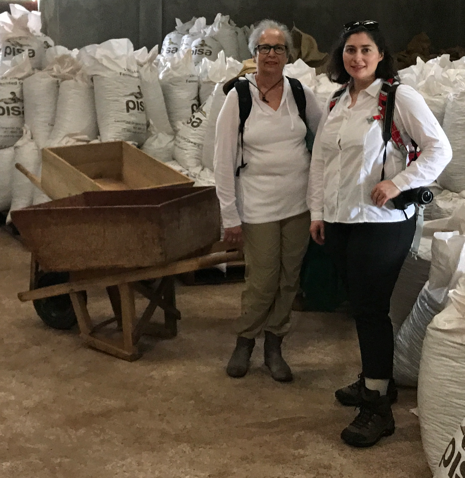 Two women wearing white tops and backpacks stand indoors in front of large white sacks and a wooden wheelbarrow. The setting appears to be industrial or agricultural.