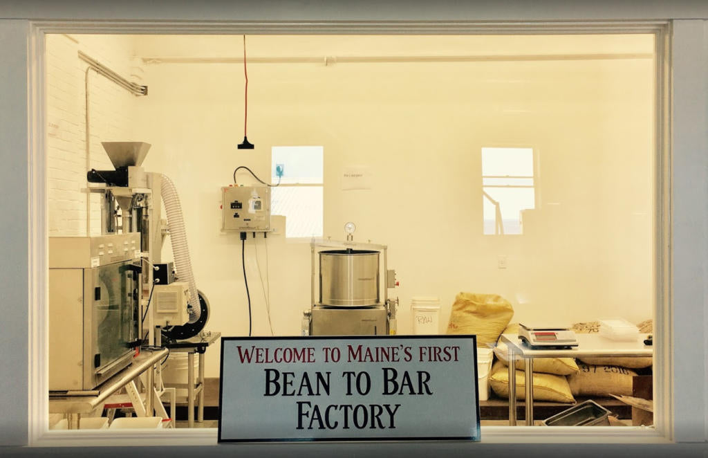 A sign reading Welcome to Maines First Bean to Bar Factory is displayed in the window of a small, clean chocolate factory with machinery and sacks of cocoa beans visible inside.