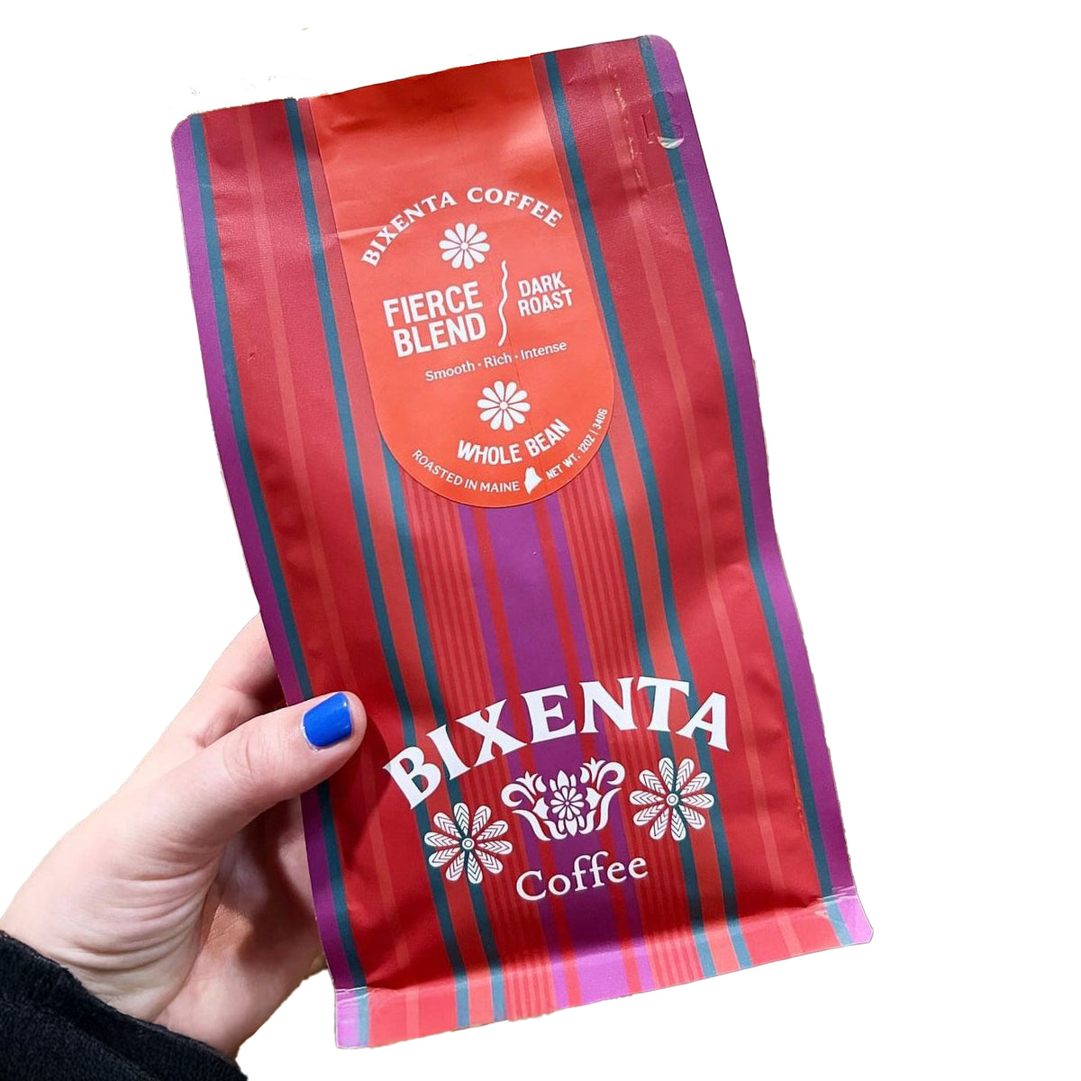 A hand with blue-painted nails holds a red bag of Bixenta Coffee by Bixby Chocolates, Fierce Blend Dark Roast whole bean. Made with Honduras Cosma beans, this small-batch coffee offers smooth, rich flavor in eye-catching packaging.