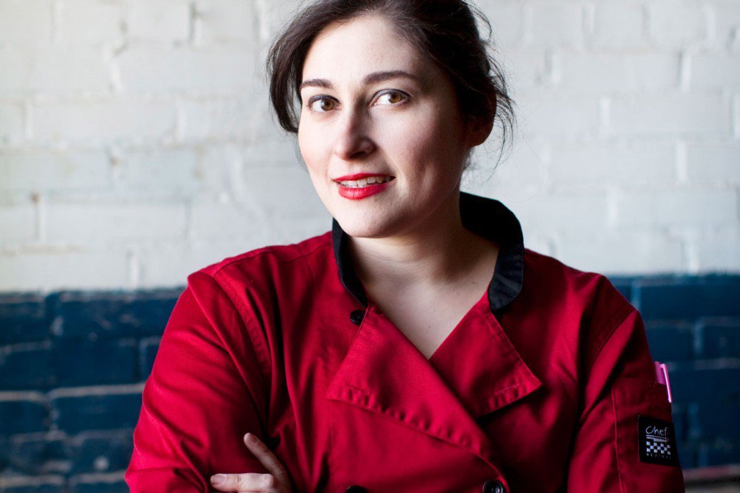 A woman wearing a red chef’s coat stands in front of a white brick wall with a blue stripe, smiling confidently with her arms crossed.