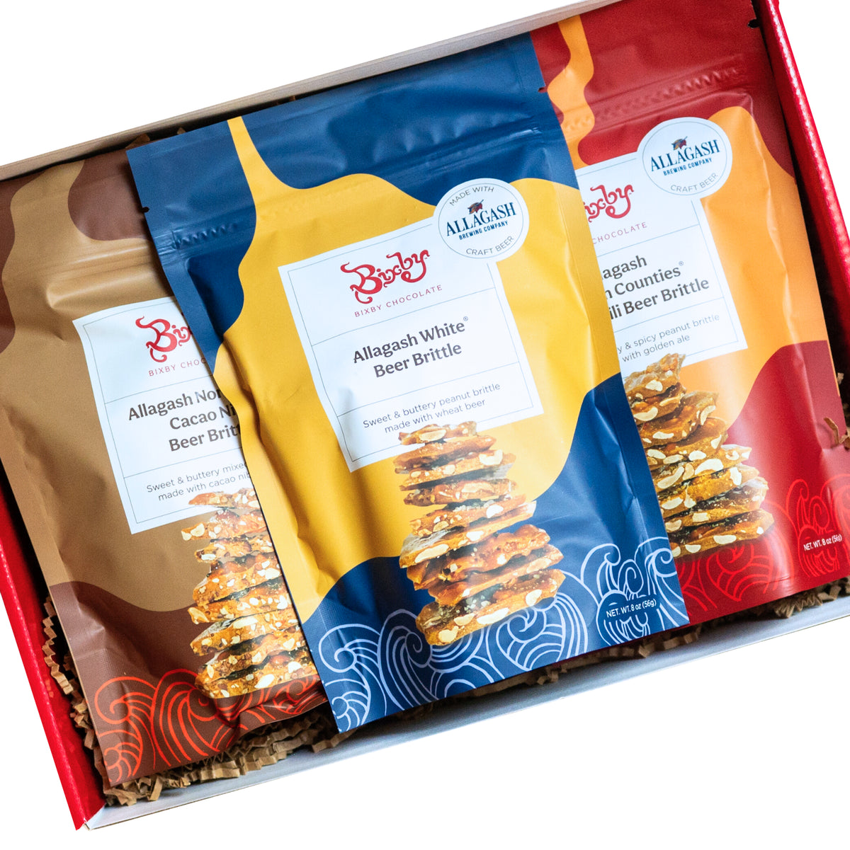 Three colorful bags of Bixby Chocolates Beer Brittle (“Allagash White,” “Cacao,” and “Four-County”) sit in a red Beer Brittle Gift Box with shredded paper, each bag showing stacks of artisanal brittle made with Allagash Beer.