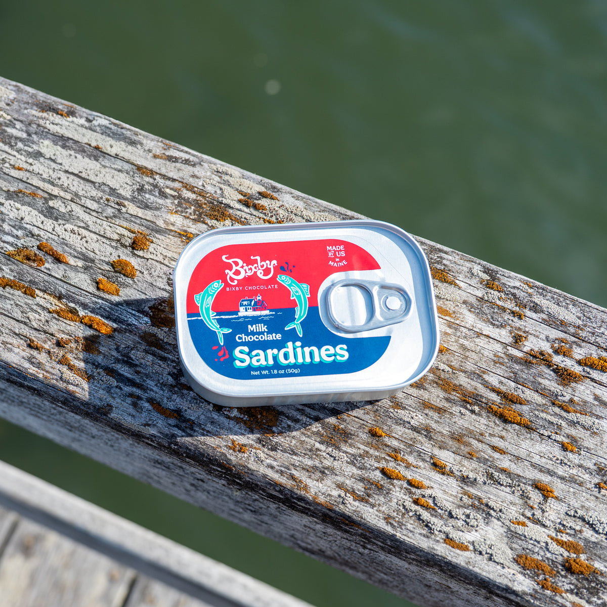 A tin of Bixby Chocolates’ Chocolate Sardines, a unique Maine souvenir, rests on a weathered wooden railing above green water, sunlight revealing orange lichen and casting shadows across the scene.