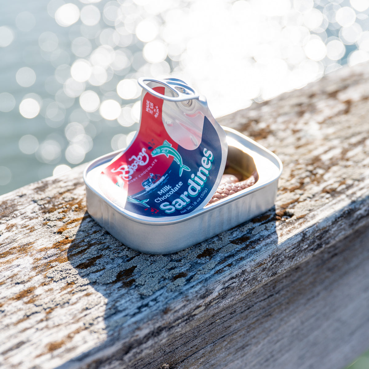 A tin of Bixby Chocolates’ Chocolate Sardines with vibrant packaging rests on a rustic wooden railing, sunlight sparkling off the water behind—a delightful Maine souvenir.