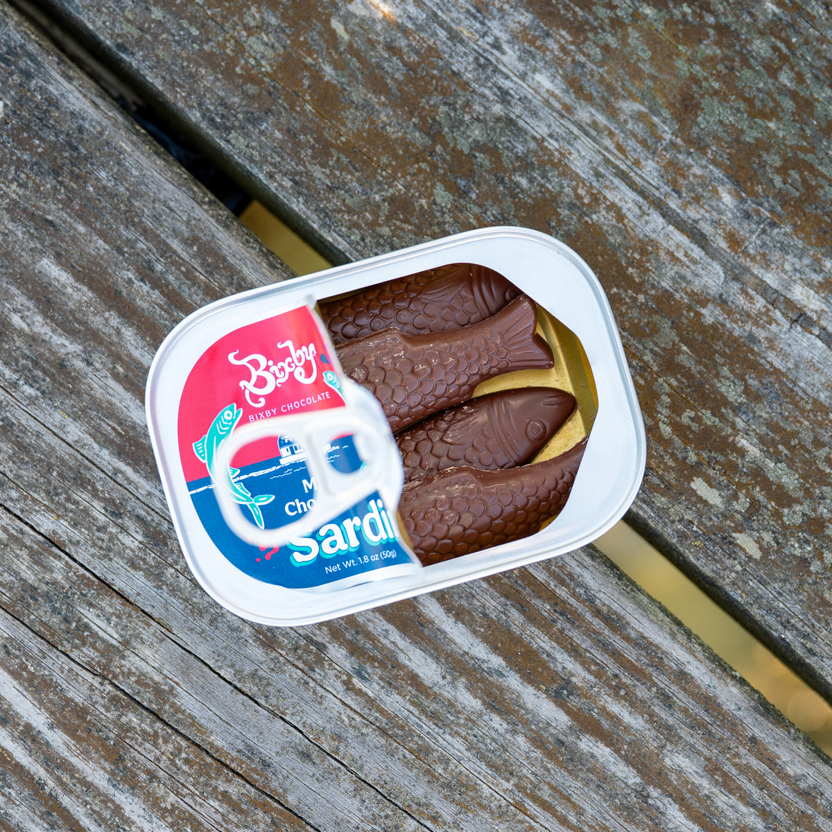 An open tin of Bixby Chocolates’ Chocolate Sardines reveals three fish-shaped artisanal chocolates inside, resembling sardines. The can rests on a weathered wood surface—a whimsical Maine souvenir.