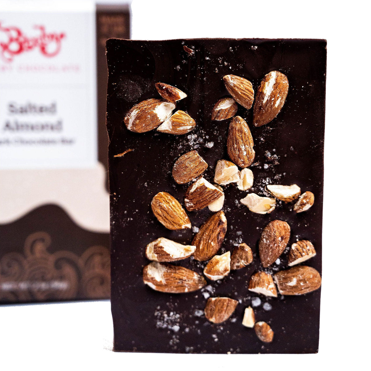 The Bixby Chocolates Dark Chocolate Salted Almond Chocolate Bar, made with 70% dark chocolate, salted almonds, and sea salt flakes, stands upright in front of a blurred product box background.