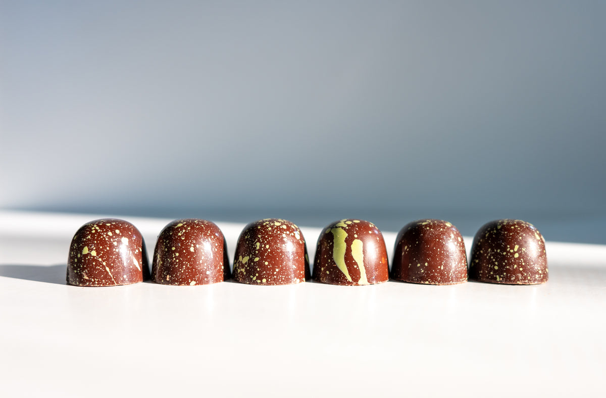 Six artisanal Pistachio Dark Chocolate Bon Bons (Vegan) from Bixby Chocolates, with gold speckles, are lined up on a white surface against a gradient light blue background, showcased in the plant-based Gift Box.