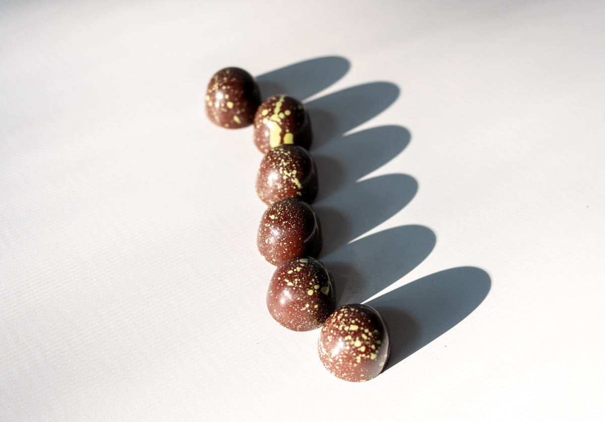 Six Pistachio Dark Chocolate Bon Bons (Vegan) by Bixby Chocolates, round and glossy with yellow speckles, are arranged diagonally on a white surface, each casting an oval shadow to the right.