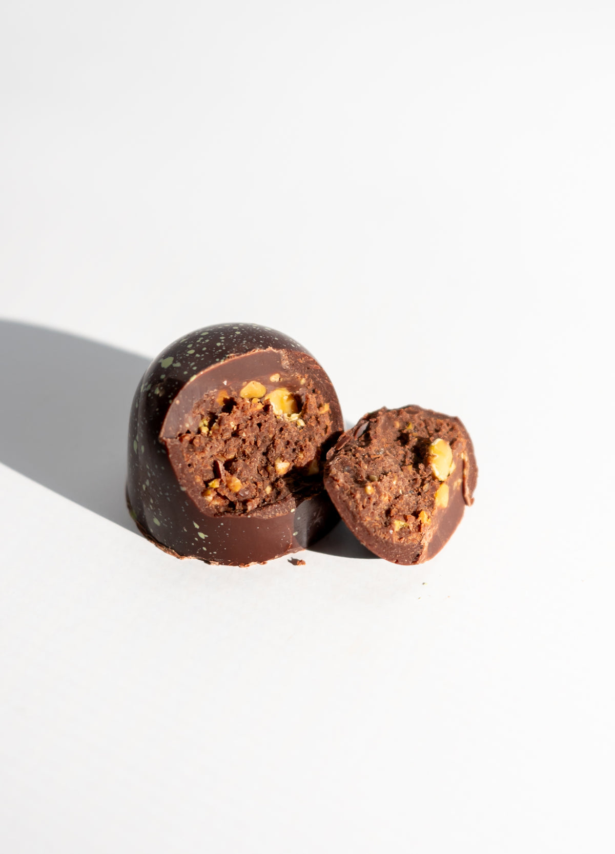 A Pistachio Dark Chocolate Bon Bon (Vegan) by Bixby Chocolates is cut in half on a white surface, displaying its crunchy pistachio gianduja filling and nutty texture. A shadow appears on the left side.
