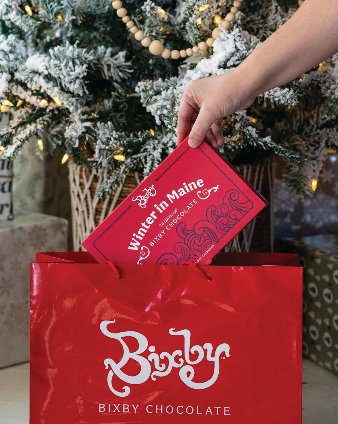 A hand places a red “Winter in Maine” Bixby Chocolate box into a matching Bixby gift bag in front of a decorated, snow-covered Christmas tree.