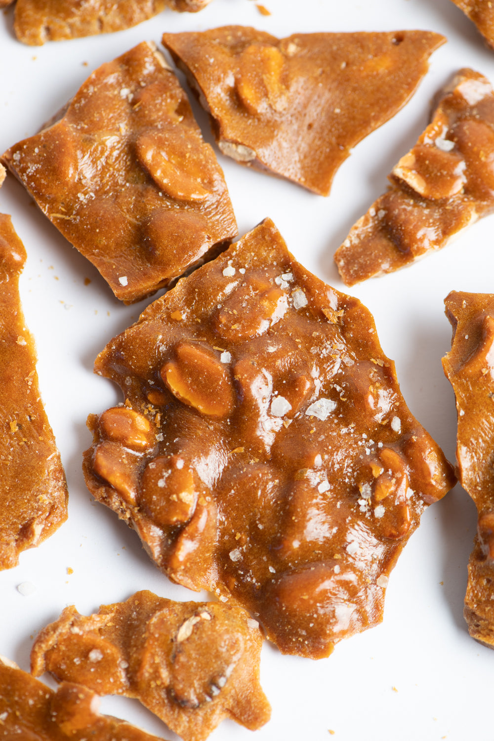 Close-up of irregularly broken pieces of golden brown peanut brittle, with visible peanuts and a glossy, caramelized texture, scattered on a white surface.
