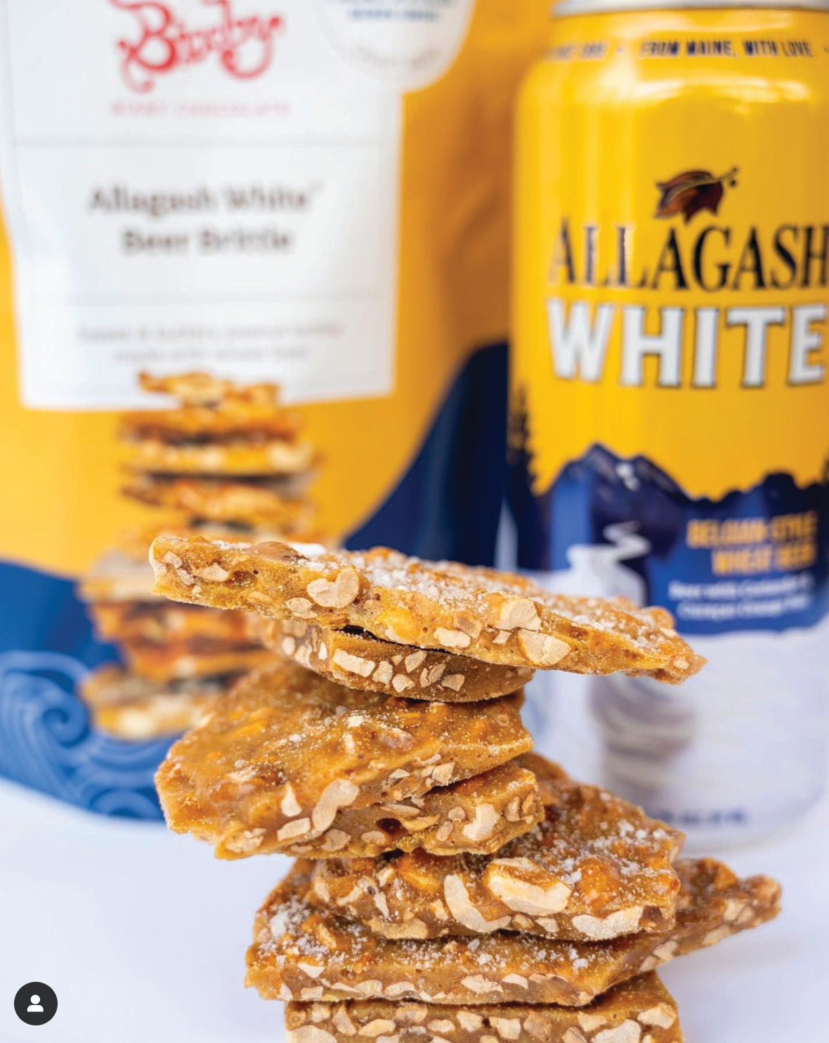 A close-up of stacked pieces of brittle with visible nuts, in front of an Allagash White beer can and a bag labeled “Allagash White Beer Brittle.” The background is slightly blurred.