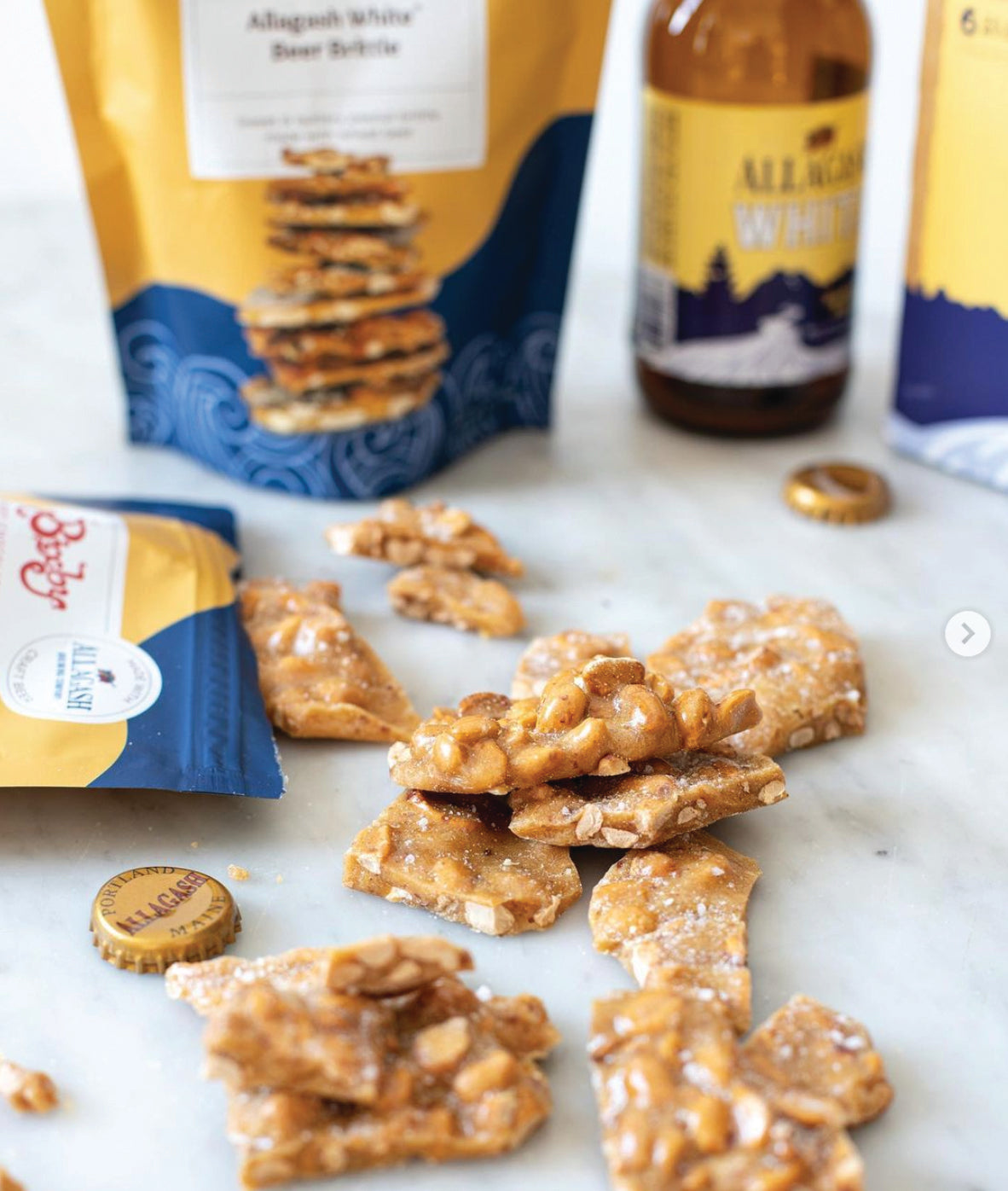 Pieces of peanut brittle are scattered on a white surface, surrounded by Allagash White beer bottles, bottle caps, and snack packaging in yellow and blue colors.
