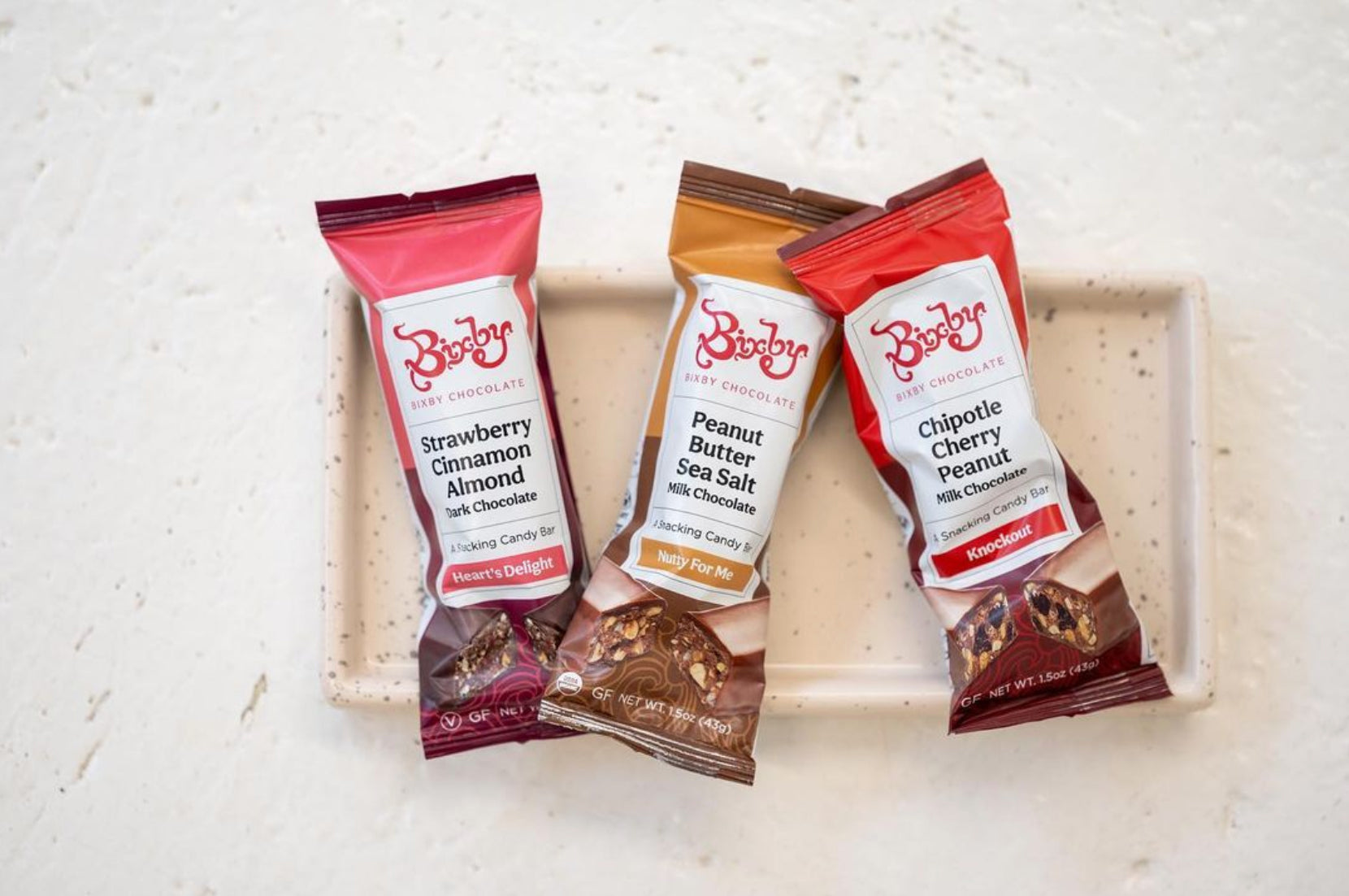 Three Bixby chocolate bars in different flavors—Strawberry Cinnamon Almond, Peanut Butter Sea Salt, and Chipotle Cherry Peanut—are arranged on a rectangular white ceramic tray on a light surface.