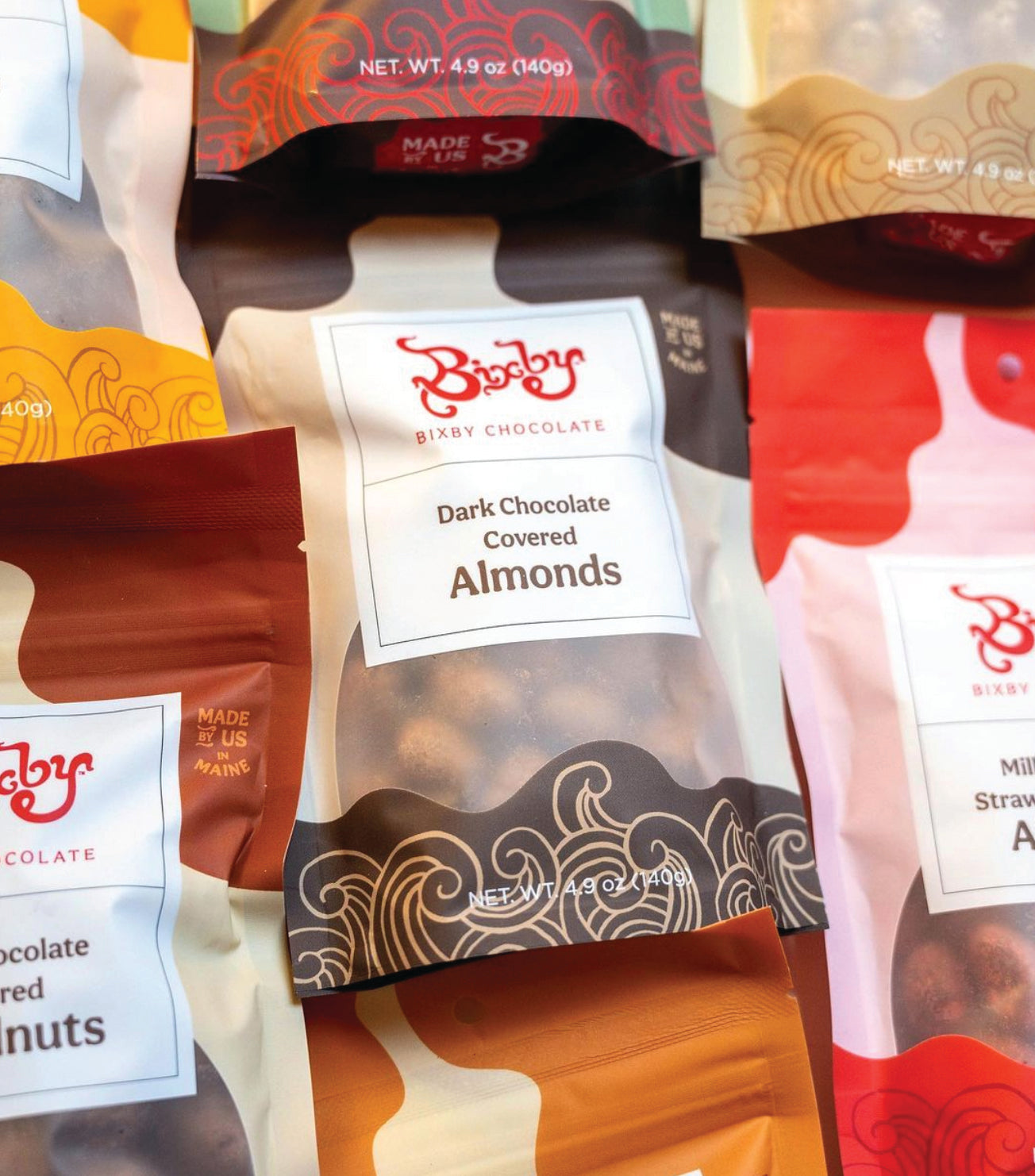 Assorted Bixby Chocolate snack bags, including Dark Chocolate Covered Almonds, arranged closely together. Each bag features a colorful, abstract design with brand logo and flavor label visible.