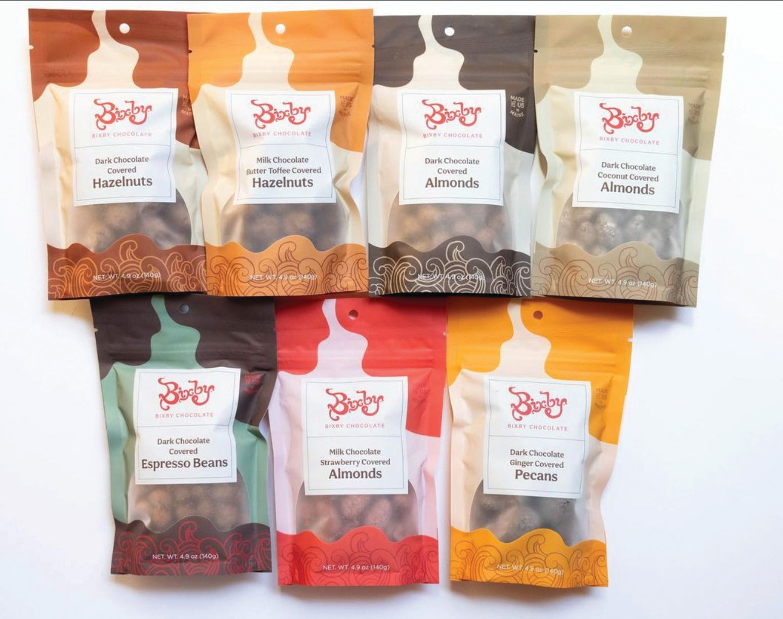 Seven bags of Bixby & Co. chocolate-covered snacks, including hazelnuts, almonds, espresso beans, and pecans, arranged in two rows on a white background. Each bag has a different color and label.