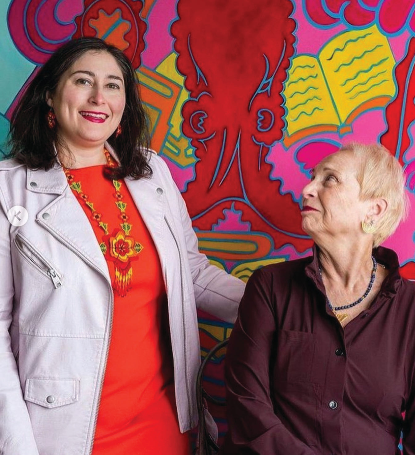 Two women pose in front of a colorful, artistic mural. The woman on the left stands and smiles, wearing a red dress and light jacket; the woman on the right sits, looking up at her, wearing a dark shirt.