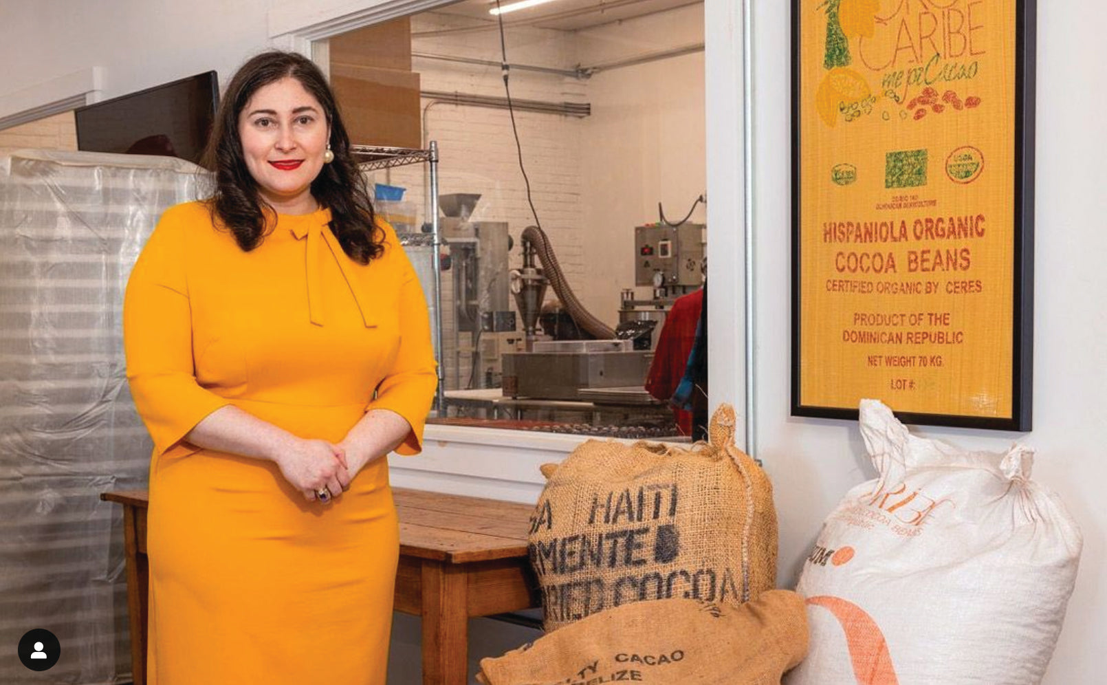 A woman in a bright orange dress stands indoors next to large sacks of cacao beans and a framed poster about Hispaniola organic cocoa beans. Industrial equipment is visible through a window behind her.
