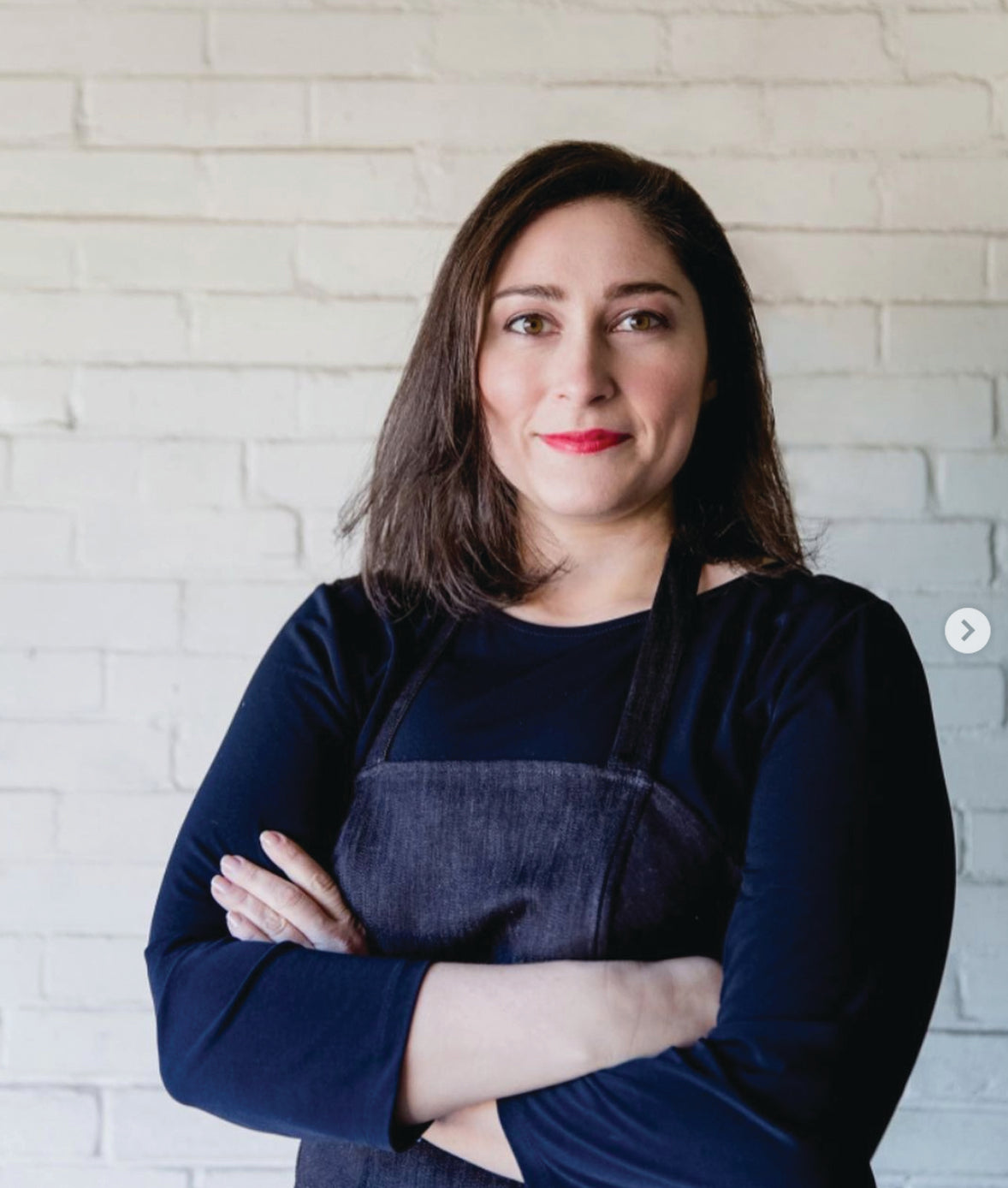 A woman with straight brown hair, wearing a black apron over a dark long-sleeve shirt, stands with her arms crossed and smiles softly in front of a white brick wall.
