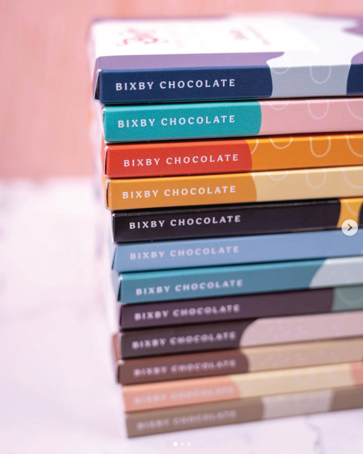 A stack of colorful Bixby Chocolate bars is arranged vertically, with each bar featuring a different pastel wrapper. The text BIXBY CHOCOLATE is printed on the side of each bar.