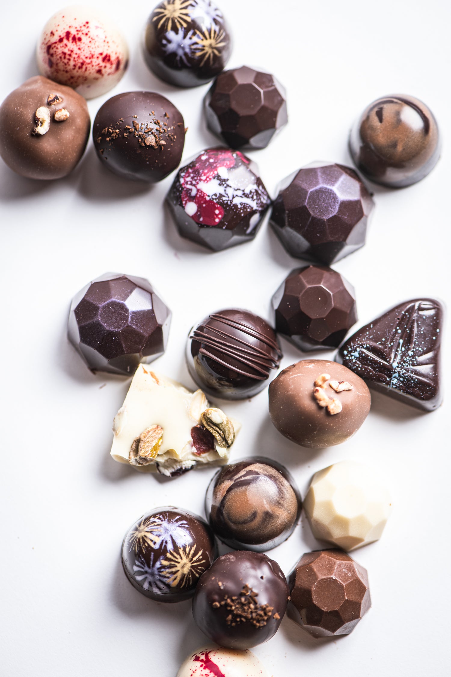 An assortment of decorative, assorted chocolates and truffles in various shapes and colors, arranged on a white background. Some are round, geometric, marbled, or topped with nuts and artistic splatters.