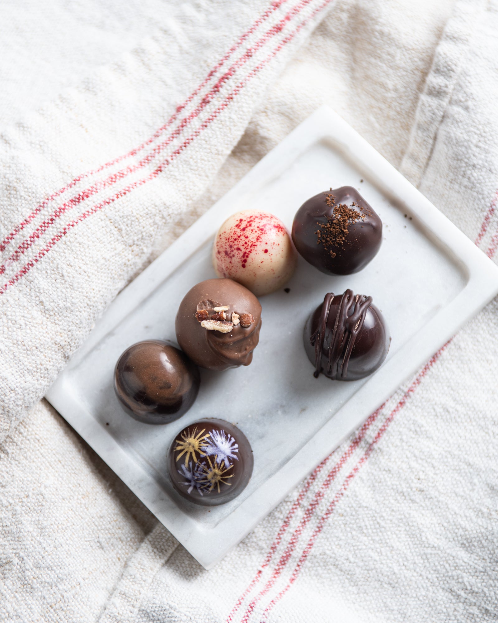 An assortment of six decorated chocolate truffles on a white marble tray, resting on a white cloth with red stripes.