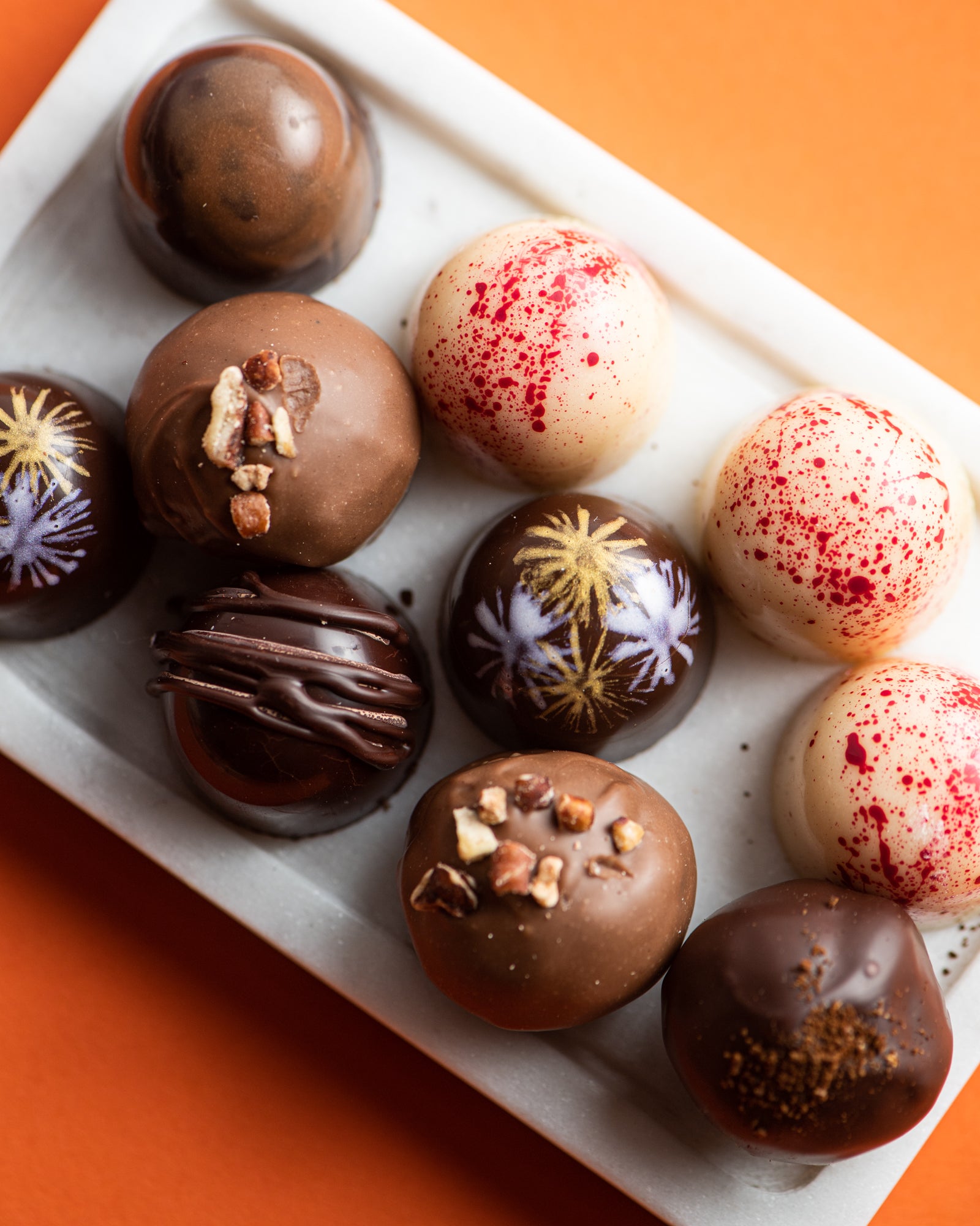 A tray of assorted chocolate truffles, some decorated with colorful patterns, nuts, or chocolate drizzle, is displayed on a white dish with an orange background.