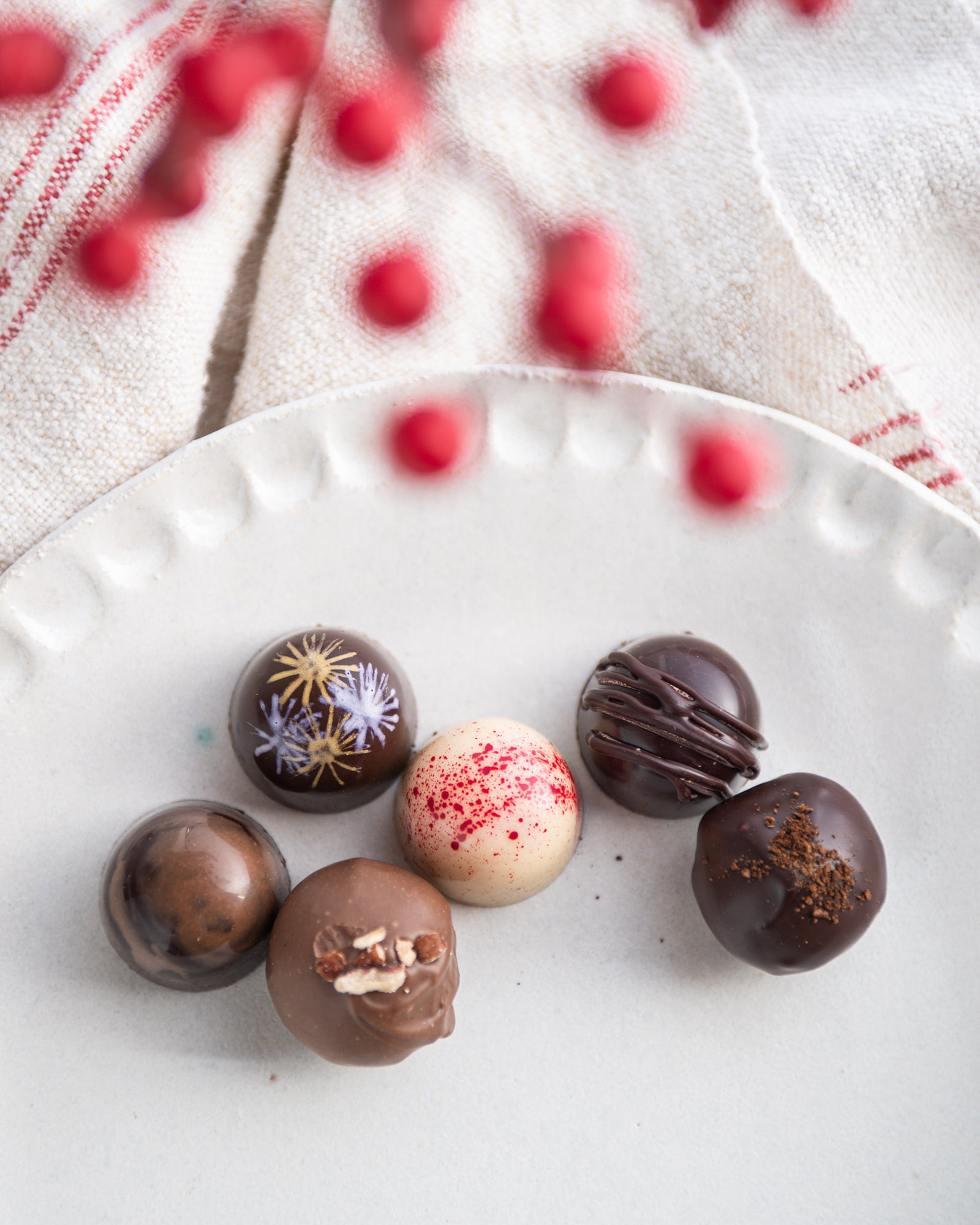 Six assorted chocolate truffles are arranged on a white plate. A cream-colored cloth and red berries are visible in the background, creating a festive and elegant atmosphere.