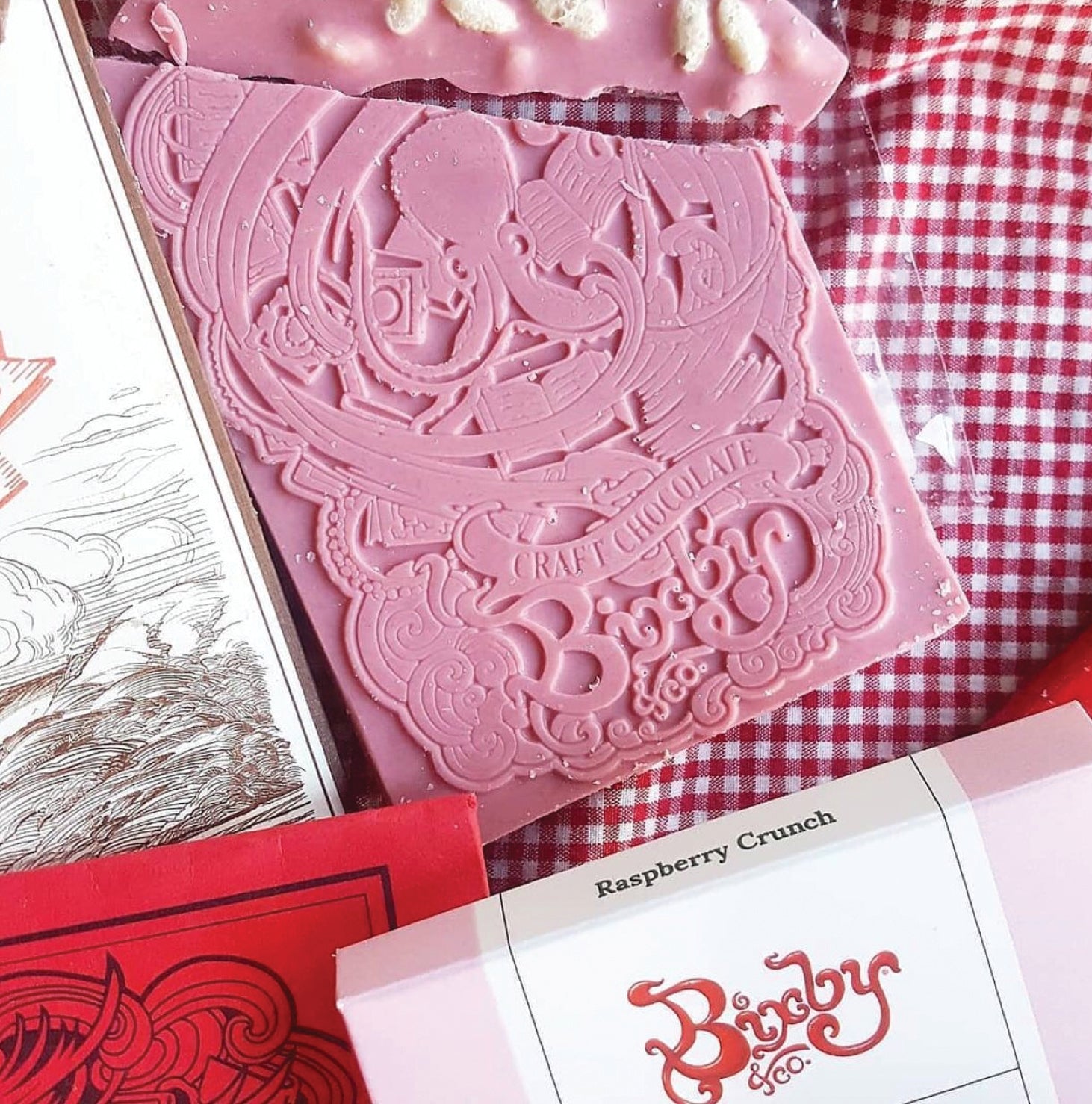 A pink chocolate bar with ornate designs and the words Craft Chocolate Bixby & Co rests on a red and white gingham cloth, near a pink box labeled Raspberry Crunch.