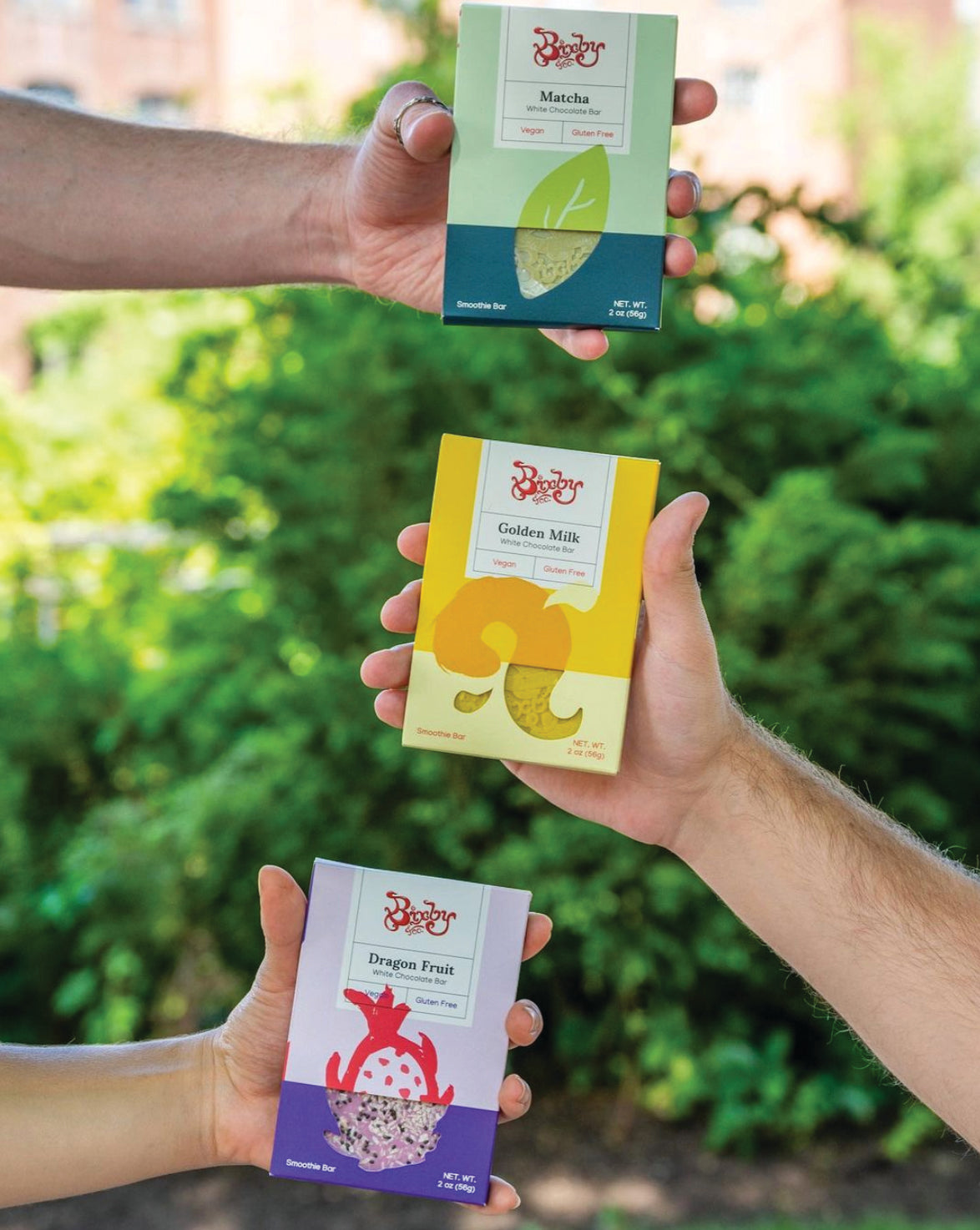 Three hands hold up colorful boxes of Byers Crackers snacks outdoors; the flavors displayed are Matcha (green box), Golden Milk (yellow box), and Dragon Fruit (purple box), with greenery in the background.
