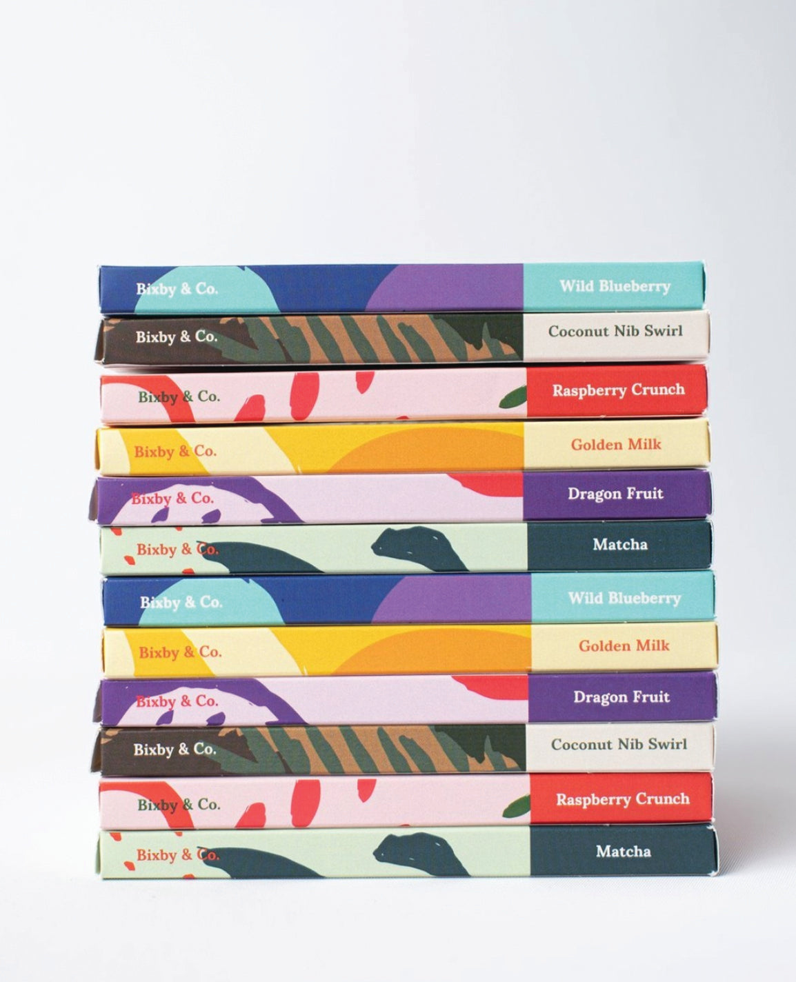 A stack of colorful chocolate bars from Bixby & Co., each with a unique patterned wrapper and labeled flavors such as Wild Blueberry, Coconut Nib Swirl, Raspberry Crunch, Golden Milk, Dragon Fruit, and Matcha.