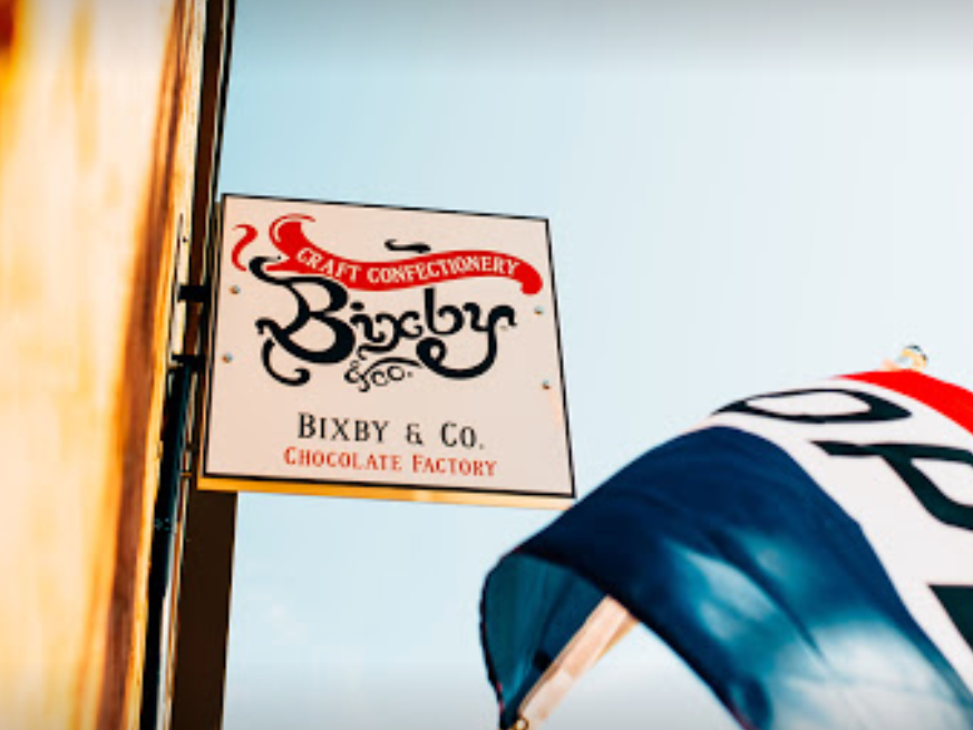 A sign for Bixby & Co. Chocolate Factory hangs on a building, reading Craft Confectionery Bixby & Co. Chocolate Factory, with part of a red, white, and blue flag visible in the lower right corner.