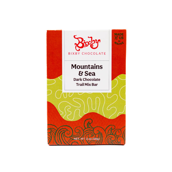 Front of a Bixby Chocolates package labeled Mountains + Sea Trail Mix 70% Dark Chocolate Bar, with a white label, red and green abstract design, net weight 2 oz (56g), and described as a wild blueberry snack and dark chocolate energy bar.