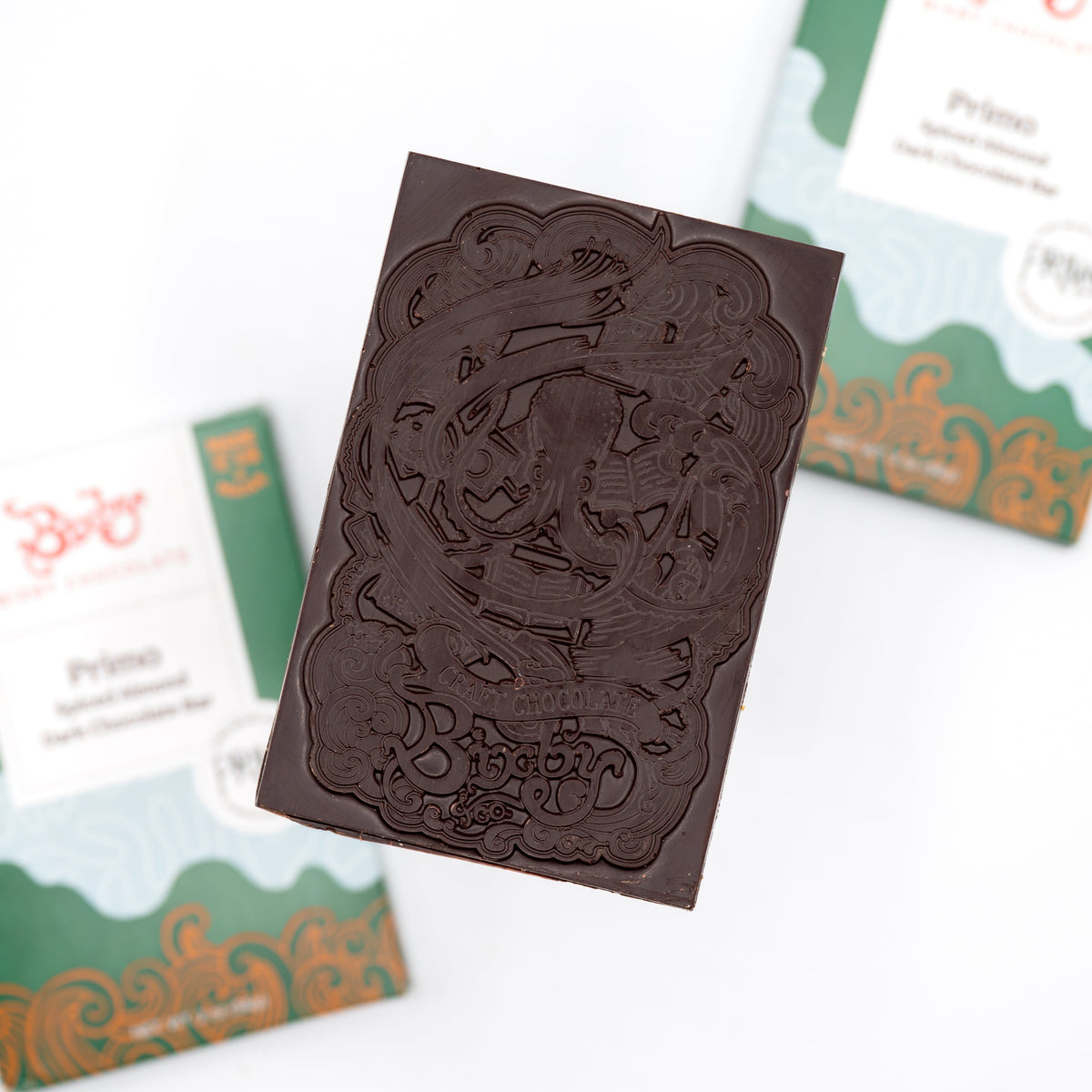 A Primo Dark Chocolate Spiced Olive Oil Roasted Almond Bar by Bixby Chocolates, crafted from Dominican Republic organic cocoa beans, features intricate designs and text above two wrappers with green, white, and gold patterns.
