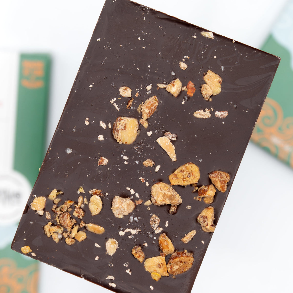 A close-up of a Bixby Chocolates Primo Dark Chocolate Spiced Olive Oil Roasted Almond Bar, topped with almonds and placed on a blurred background with green and white packaging.