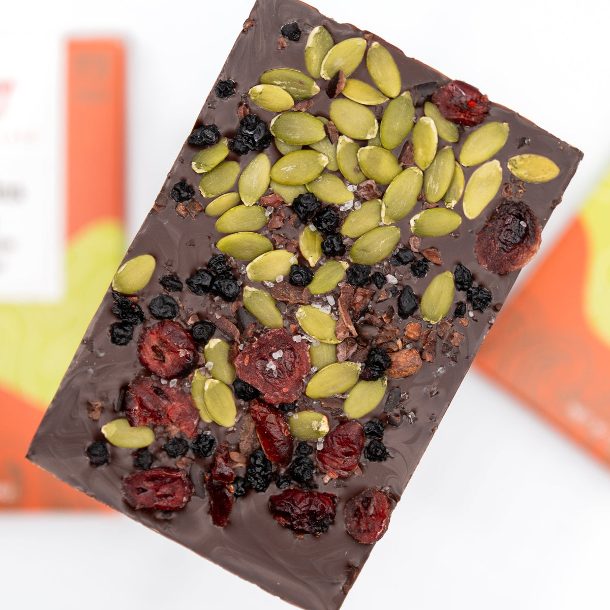 The Mountains + Sea Trail Mix 70% Dark Chocolate Bar by Bixby Chocolates is topped with pumpkin seeds, dried cranberries, blueberries, and cacao nibs, set against a blurred orange and white background.