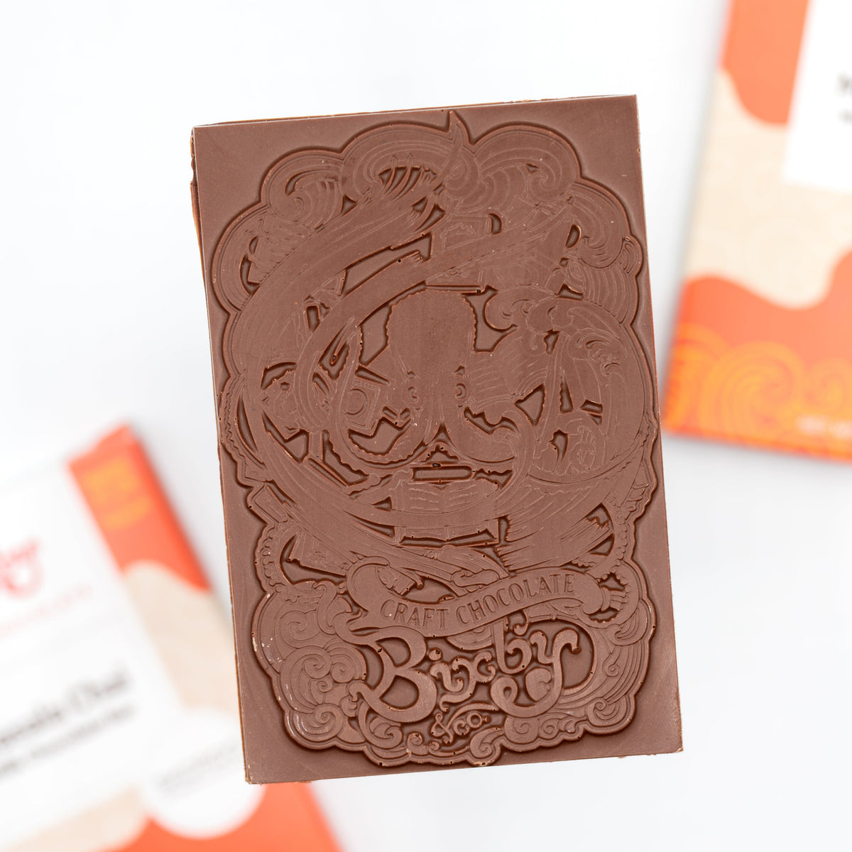 A rectangular Bixby Chocolates bar engraved with "Craft Chocolate" and "Bixby," featuring their Skordo Chai Milk Chocolate Bar, is held above two blurred chocolate wrappers in the background.