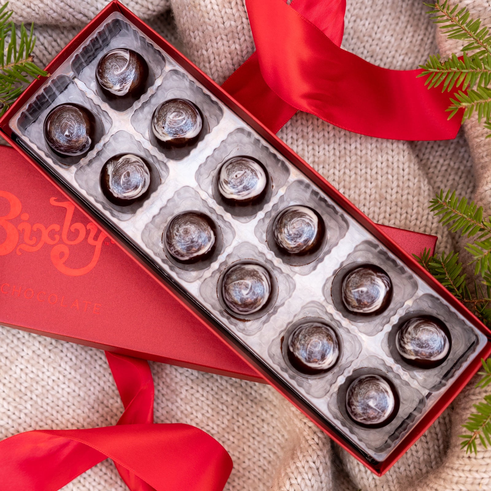 A festive red box of Bixby Chocolates Eggnog Bon Bons, open to display twelve round treats in a tray, sits on beige knit fabric with a red ribbon and pine—perfect for savoring these holiday chocolates.