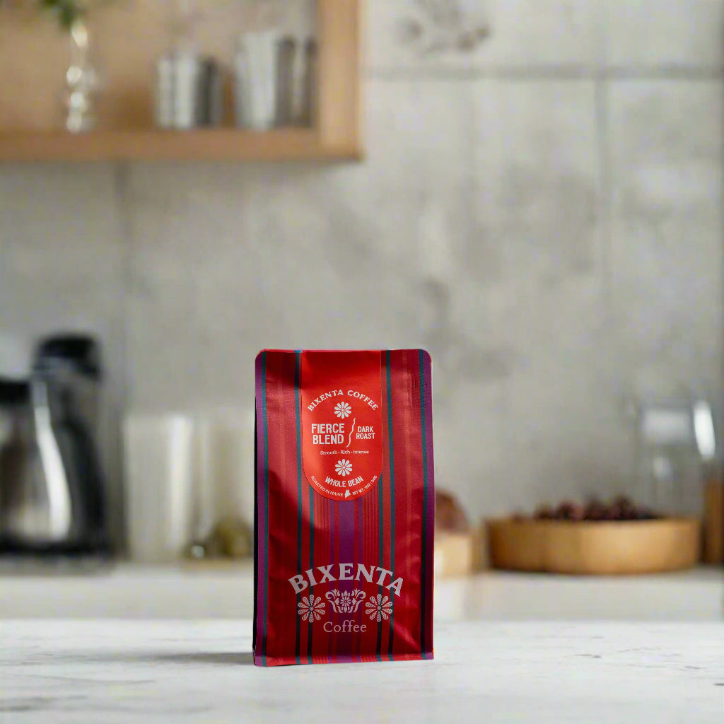 A red and purple striped bag of Bixenta Coffee by Bixby Chocolates, labeled Fierce Blend and featuring small-batch roasted Honduras Cosma beans, stands on a white countertop in a modern kitchen with blurred shelves in the background.